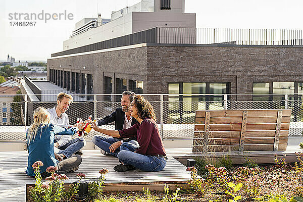 Fröhliche Kollegen stoßen beim Sitzen auf der Terrasse auf eine Flasche