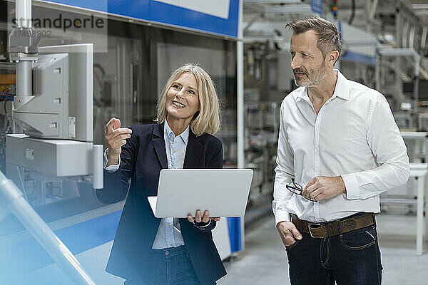Lächelnde berufstätige Frau gestikuliert  während sie mit einem männlichen Kollegen über Maschinen in der Fabrik diskutiert