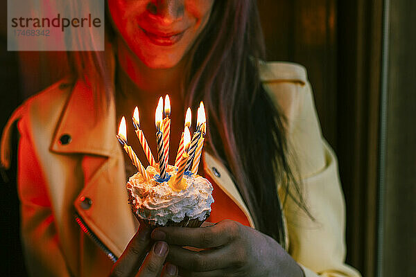 Lächelnde Frau hält Cupcake mit brennenden Kerzen am Geburtstag zu Hause