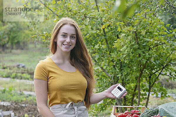 Lächelnde Bäuerin hält Kreditkartenlesegerät im Garten