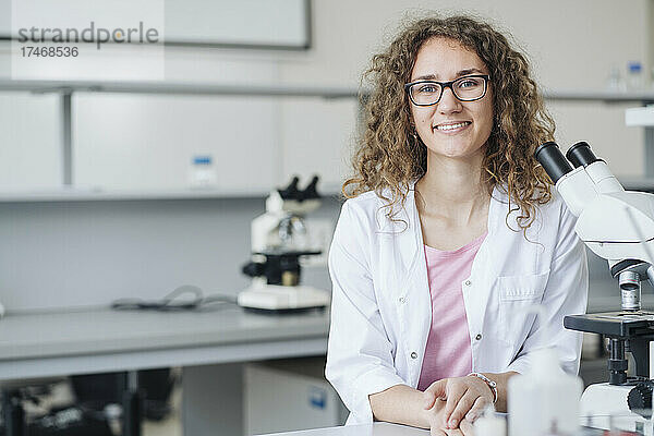 Lächelnder junger Wissenschaftler mit Mikroskop am Labortisch