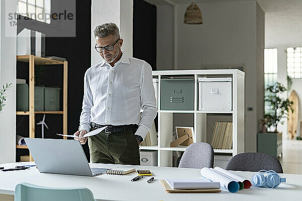 Geschäftsmann hält Dokument in der Hand  während er im Büro auf den Laptop schaut