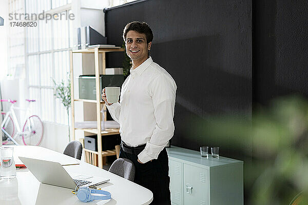Lächelnder Geschäftsmann mit der Hand in der Tasche beim Kaffeetrinken im Büro