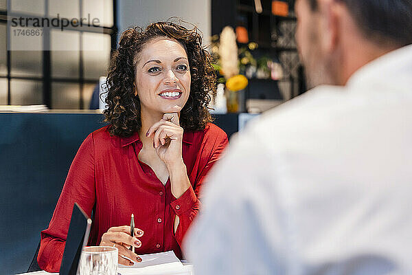 Lächelnde Geschäftsfrau mit der Hand am Kinn  die ihrem Kollegen im Büro zuhört