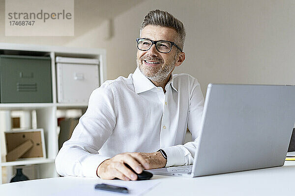 Lächelnder Geschäftsmann mit Laptop  der im Büro nachdenkt