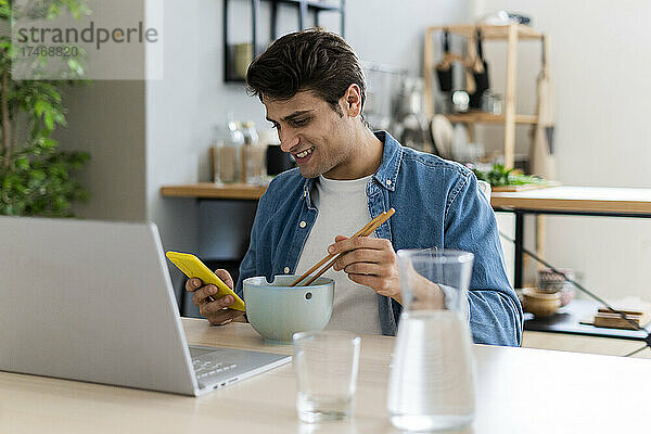 Lächelnder Mann benutzt Mobiltelefon  während er zu Hause etwas isst