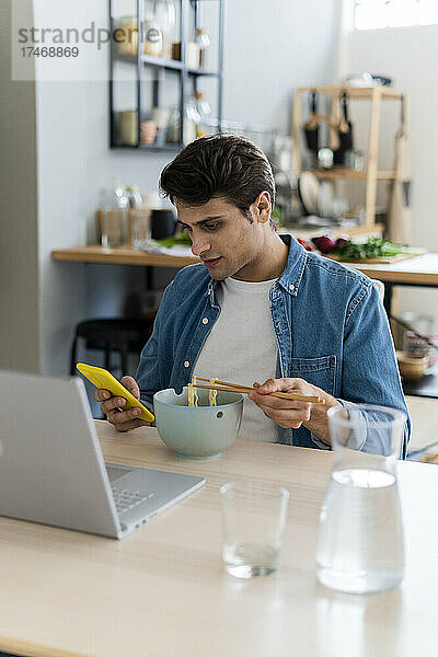 Junger Mann benutzt Smartphone  während er zu Hause Nudeln isst
