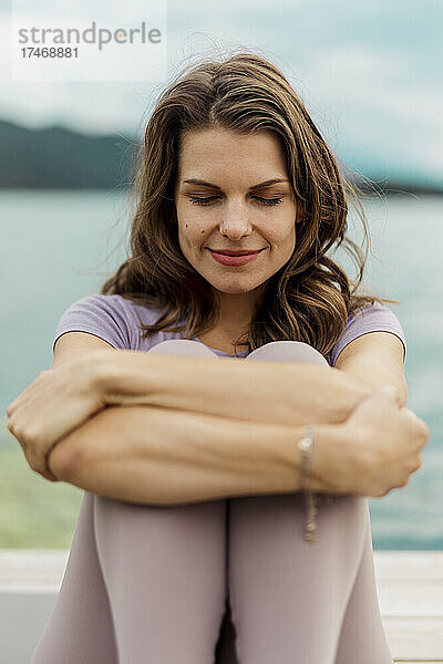 Frau mit geschlossenen Augen umarmt Knie vor dem See