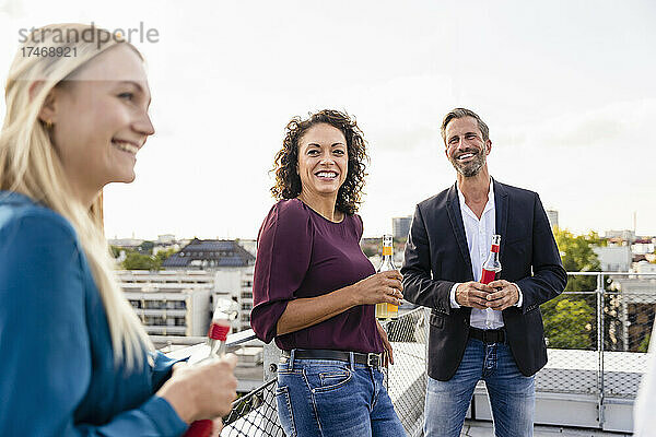 Lächelnde Geschäftsfrau hält Alkoholflasche mit Kollegen auf dem Dach