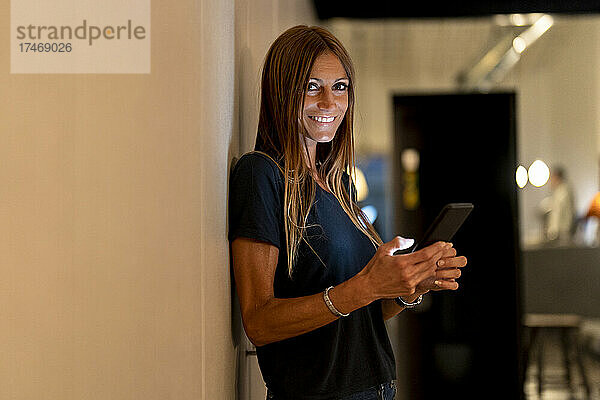 Lächelnde Frau mit Smartphone  die zu Hause an der Wand steht