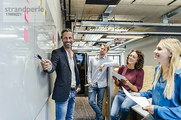 Lächelnder Geschäftsmann zeigt bei Treffen mit Kollegen im Büro auf Whiteboard