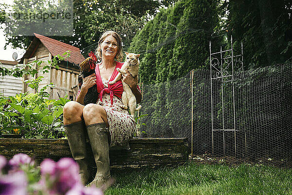Glückliche Frau trägt Katze und Hahn  während sie im Hinterhof sitzt