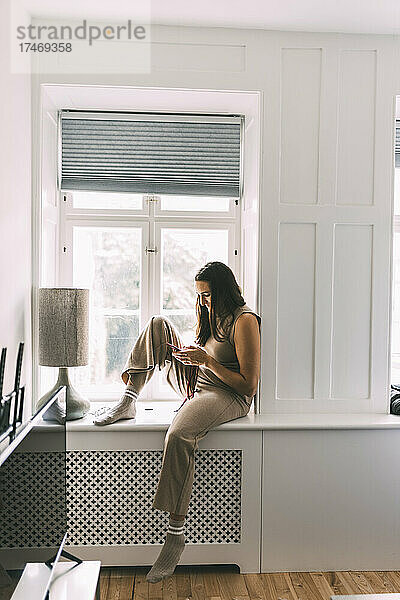 Frau sitzt zu Hause auf Fensterbank und benutzt Smartphone