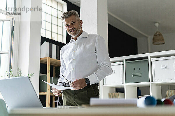 Lächelnder Geschäftsmann mit Dokument und Brille am Schreibtisch