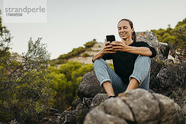 Lächelnde Frau  die ihr Mobiltelefon auf einem Felsen benutzt