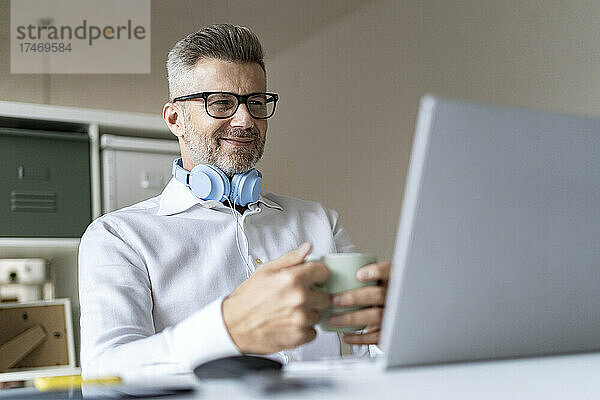 Lächelnder Geschäftsmann mit Kaffeetasse bei Videokonferenz über Laptop im Büro