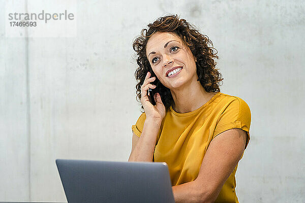 Lächelnde Geschäftsfrau mit Laptop-Hand am Kinn vor der Wand