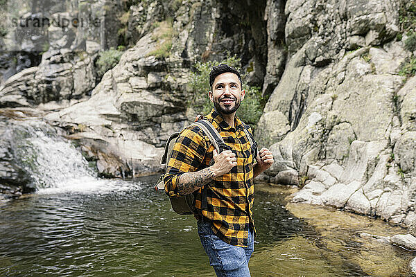 Lächelnder erwachsener Mann mit Rucksack steht am Teich im Wald