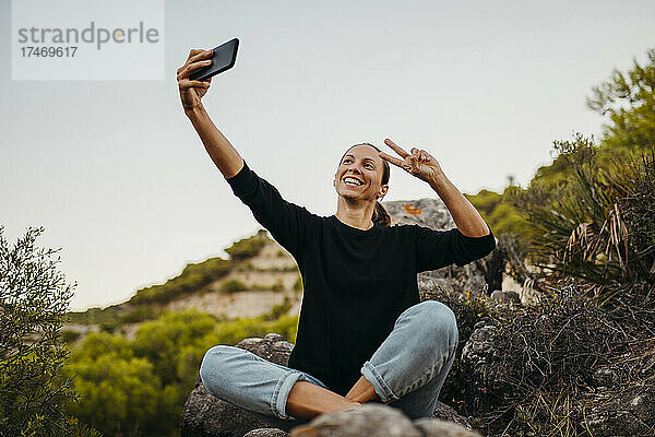 Lächelnde Frau gestikuliert  während sie ein Selfie mit ihrem Smartphone auf einem Felsen macht