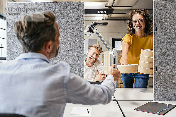 Lächelnde Geschäftsfrau gibt einem Kollegen am Schreibtisch eine Lunchbox