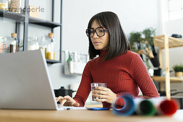 Geschäftsfrau arbeitet am Laptop und hält Wasserglas im Heimbüro