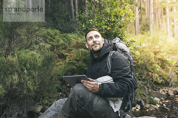 Lächelnder Mann sitzt mit digitalem Tablet im Wald