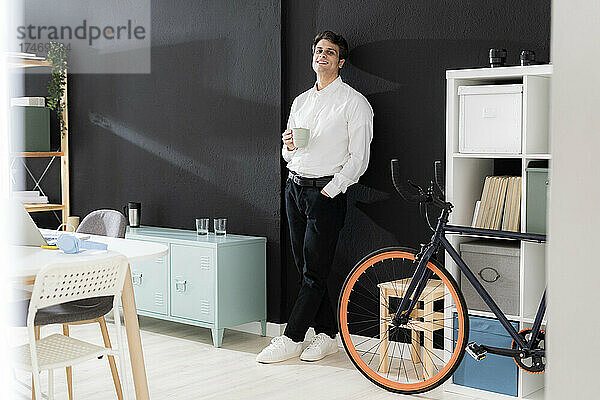 Junger Geschäftsmann mit Kaffeetasse lehnt an schwarzer Wand im Kreativbüro