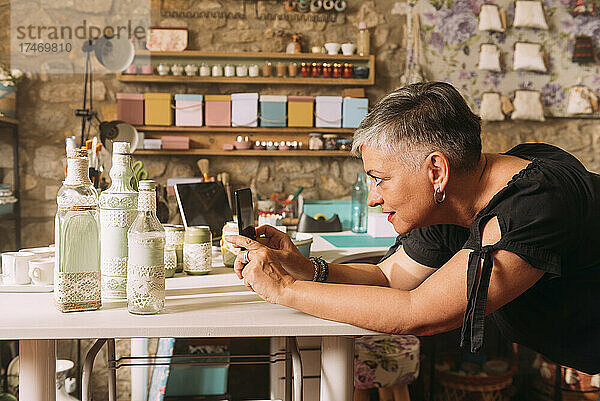 Reife Frau fotografiert Handwerksprodukte mit Smartphone an der Werkbank