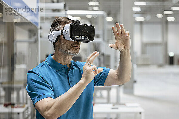 Reifer männlicher Profi gestikuliert  während er in der Fabrik einen Virtual-Reality-Simulator verwendet