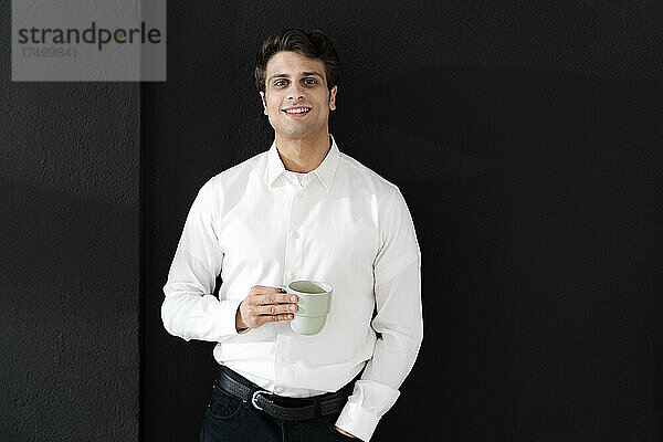 Selbstbewusster Geschäftsmann mit Kaffeetasse steht vor schwarzer Wand