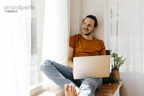 Lächelnder Mann mit Laptop sitzt am Fenster