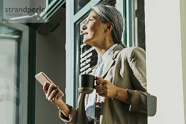 Reife Frau hält Kaffeetasse und Smartphone am Eingang des Cafés