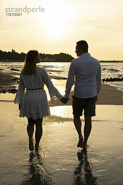 Freund und Freundin halten Händchen beim Spaziergang am Strand bei Sonnenuntergang