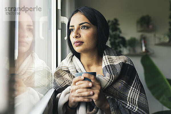 Junge Frau mit Kaffeetasse schaut zu Hause durch das Fenster