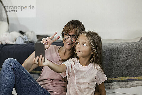 Mädchen macht Selfie mit Mutter  die zu Hause per Handy ein Friedenszeichen zeigt