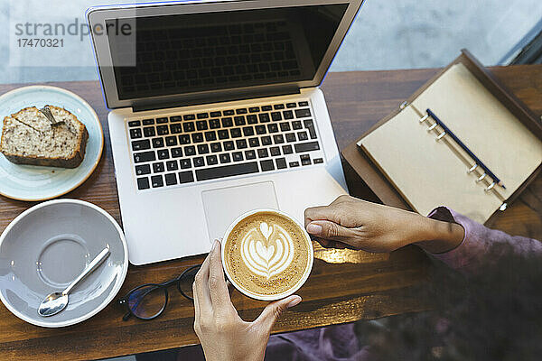 Geschäftsfrau mit Laptop hält Cappuccino-Tasse am Cafétisch