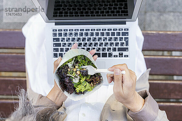 Frau mit Laptop isst Salat  während sie auf Bank sitzt