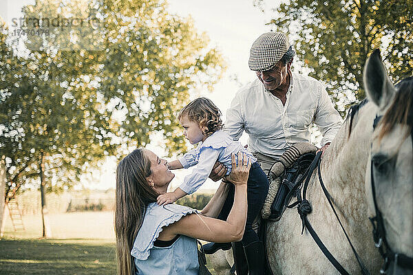 Mutter trägt Tochter  während älterer Mann auf Pferd sitzt