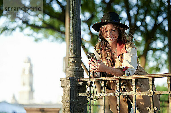 Lächelnde Frau mit Smartphone lehnt am Geländer
