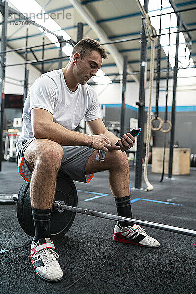 Männlicher Athlet benutzt Smartphone  während er im Fitnessstudio auf der Langhantel sitzt