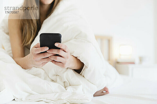 Junge Frau benutzt Mobiltelefon  während sie im Schlafzimmer auf dem Bett sitzt