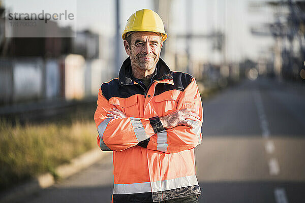 Lächelnder männlicher Hafenarbeiter mit verschränkten Armen auf der Straße in der Industrie