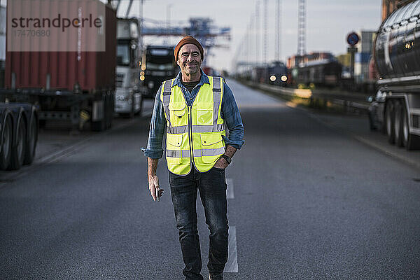 Lächelnder männlicher LKW-Fahrer mit der Hand in der Tasche  der am Dock auf der Straße läuft
