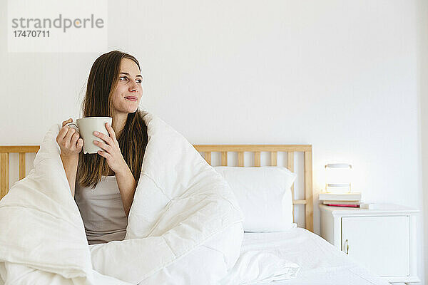 Junge Frau mit braunen Haaren trinkt Kaffee zu Hause im Bett