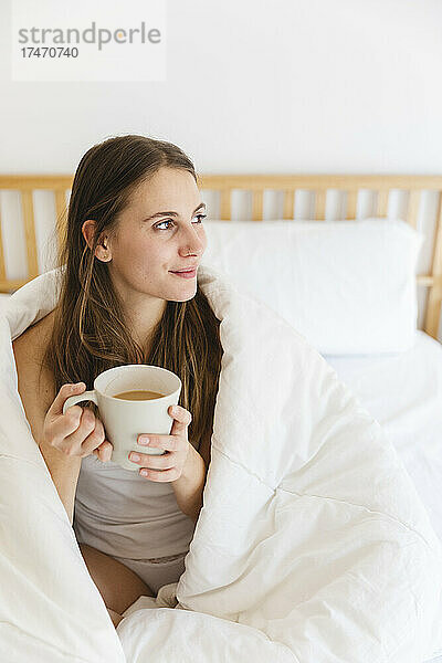 Junge Frau hält Kaffeetasse in der Hand  während sie zu Hause in eine weiße Decke gehüllt ist