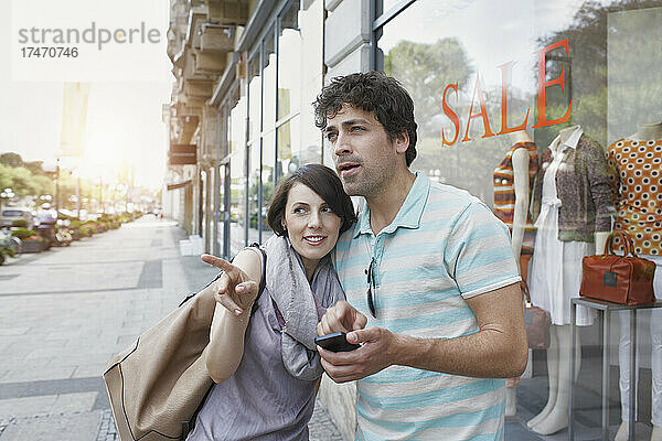 Frau zeigt  während Freund mit Smartphone neben Geschäft auf Fußweg steht