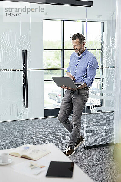 Männlicher Berufstätiger benutzt Laptop  während er sich im Büro an die Glastür lehnt