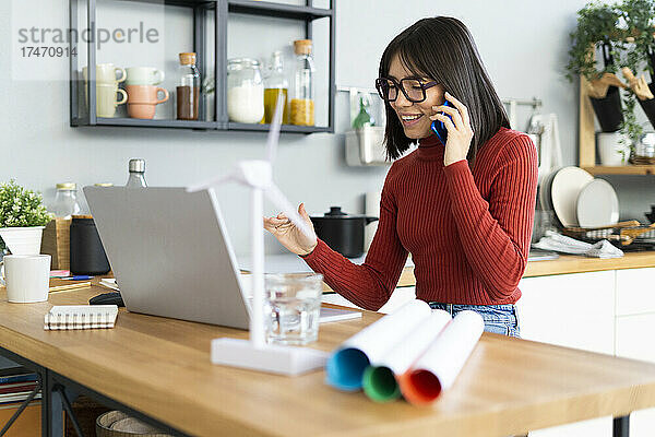 Lächelnder Freiberufler mit Laptop  der im Heimbüro mit dem Mobiltelefon telefoniert