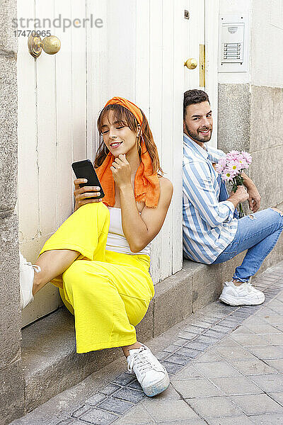 Lächelnde Freundin benutzt Mobiltelefon  während sie mit Mann an der Tür sitzt