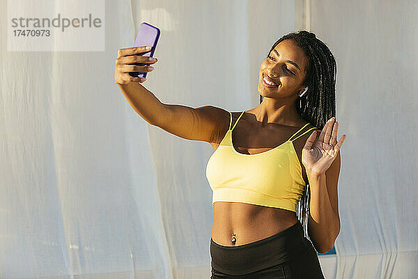 Glückliche Sportlerin winkt während eines Videoanrufs über das Mobiltelefon mit der Hand
